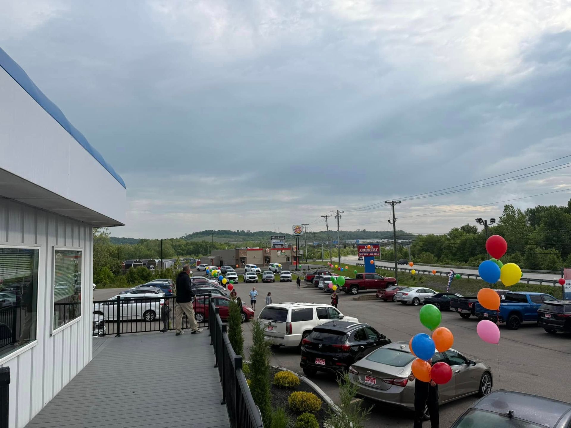 Car dealership lot with rows of cars, colorful balloons, and a cloudy sky on a rainy day
