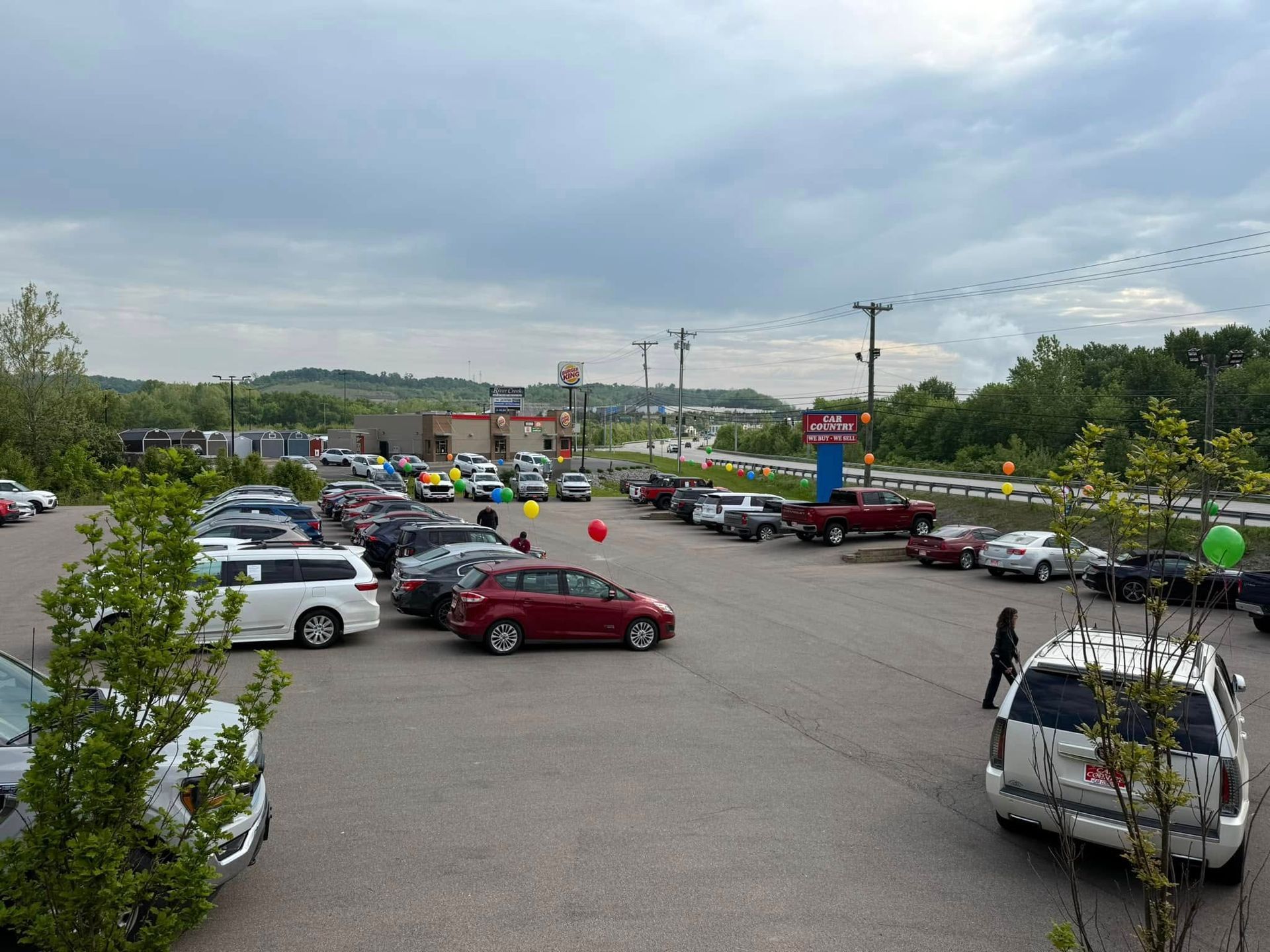 Busy parking lot with many cars, storefronts in the distance, and cloudy sky overhead