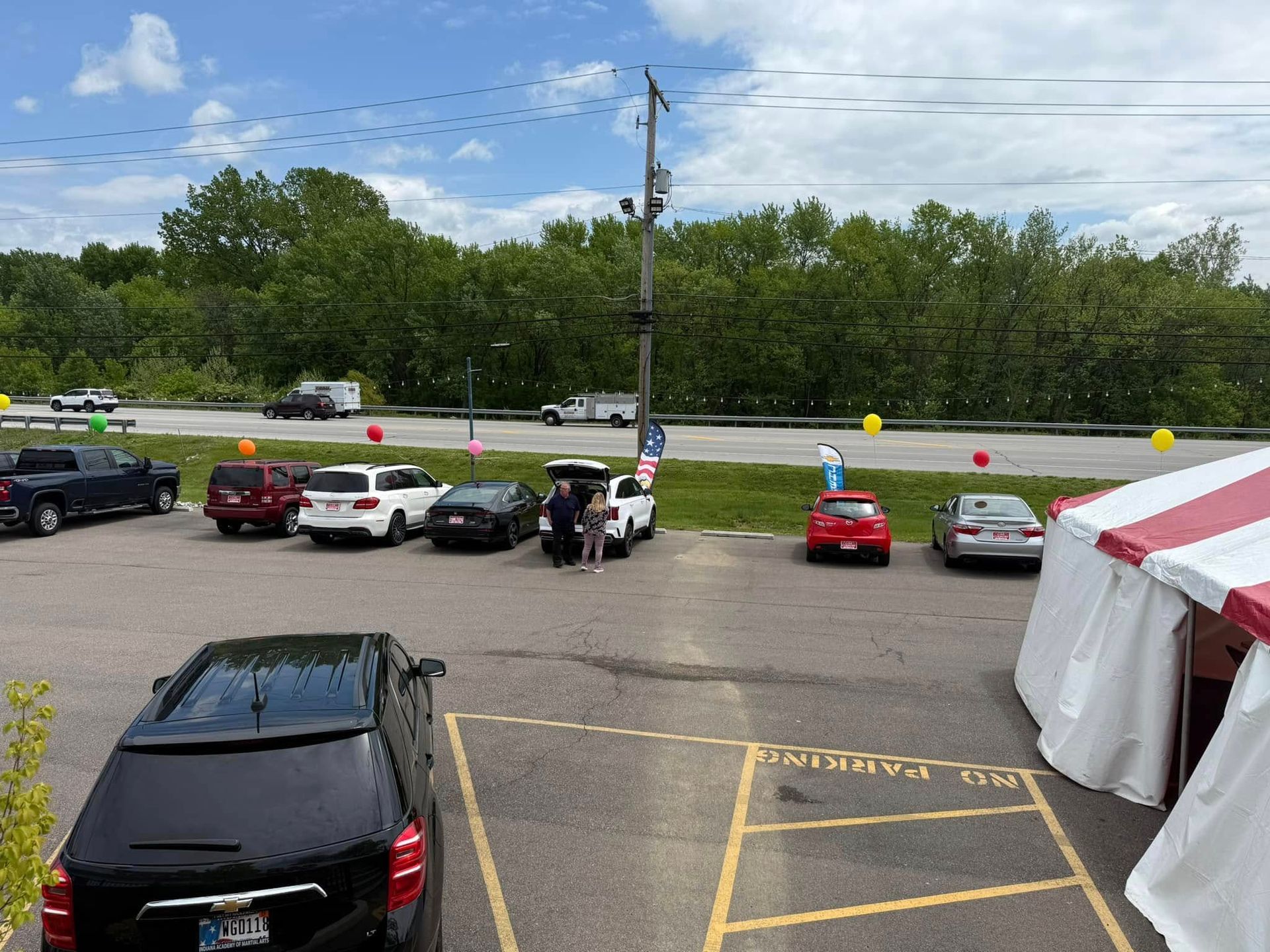 Parking lot with cars, a white tent, and several people near a road under a cloudy sky