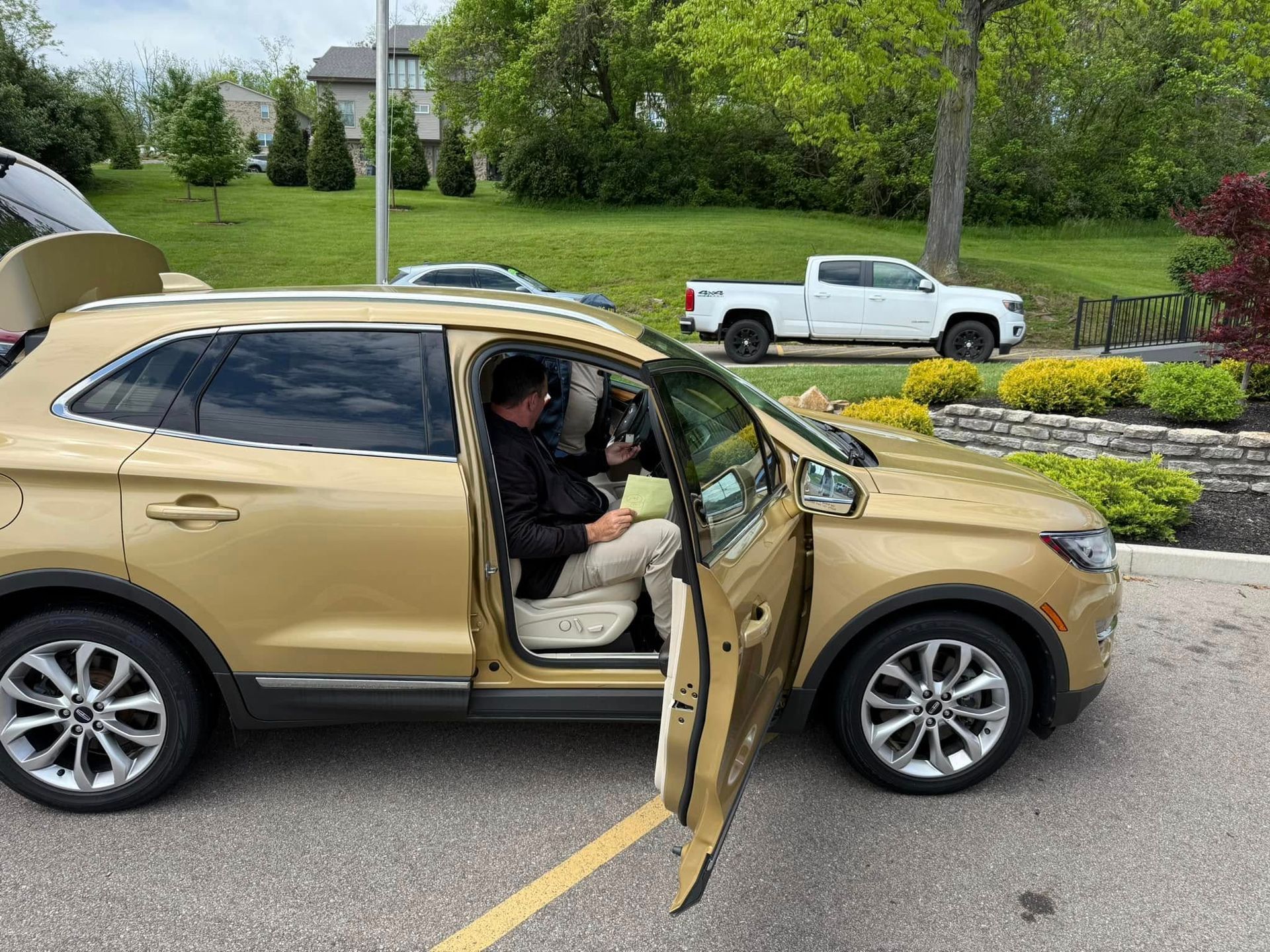 Person exiting a gold SUV parked on a driveway, with a white pickup truck and greenery in the background