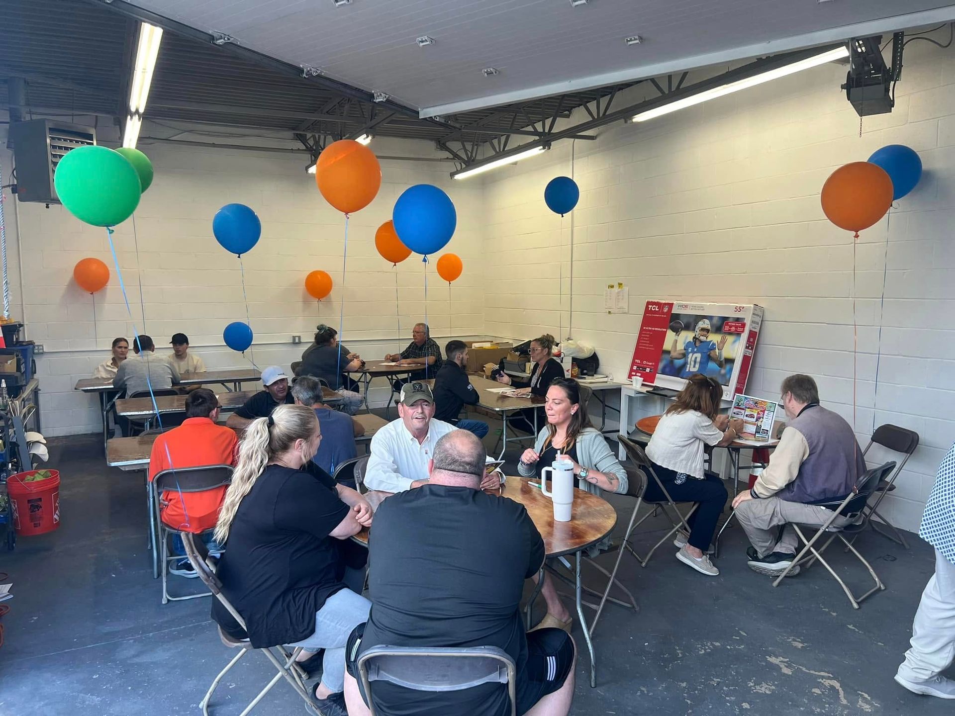 People seated at tables in a bright room with colorful balloons hanging from the ceiling.