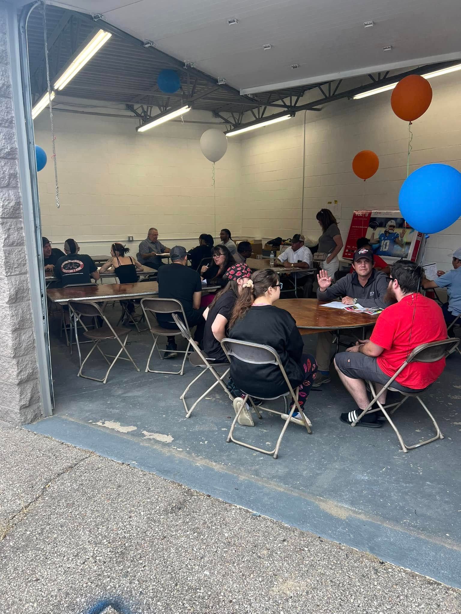 People seated at tables in a garage-like room with balloons and an American flag, social gathering inside.