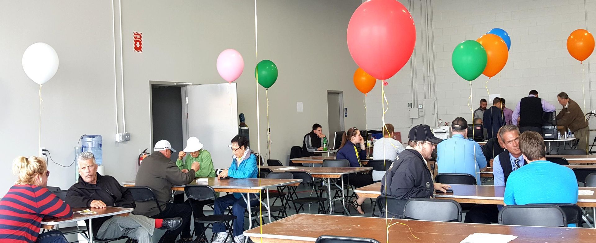 People seated at tables in a bright room decorated with colorful balloons during an indoor gathering