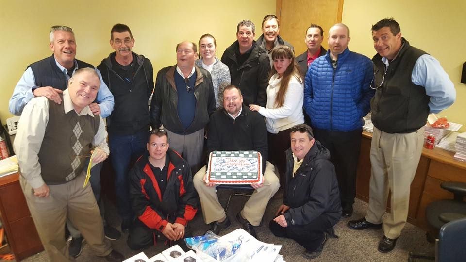 Group of people posing indoors around a cake, smiling for a celebration photo.