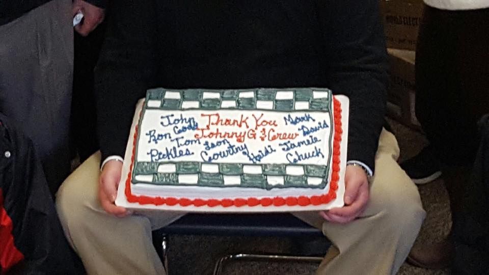 A decorated rectangular cake with handwritten icing, held in someone’s lap on a chair.
