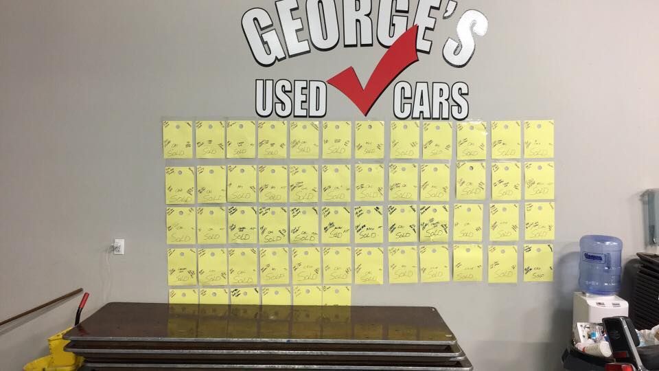 Wall of yellow sticky notes beneath a George’s Used Cars sign in a waiting area