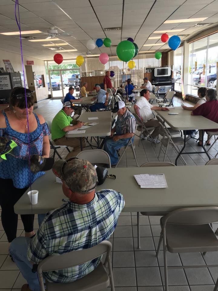 Community gathering in a bright hall with people seated at folding tables and colorful balloons overhead