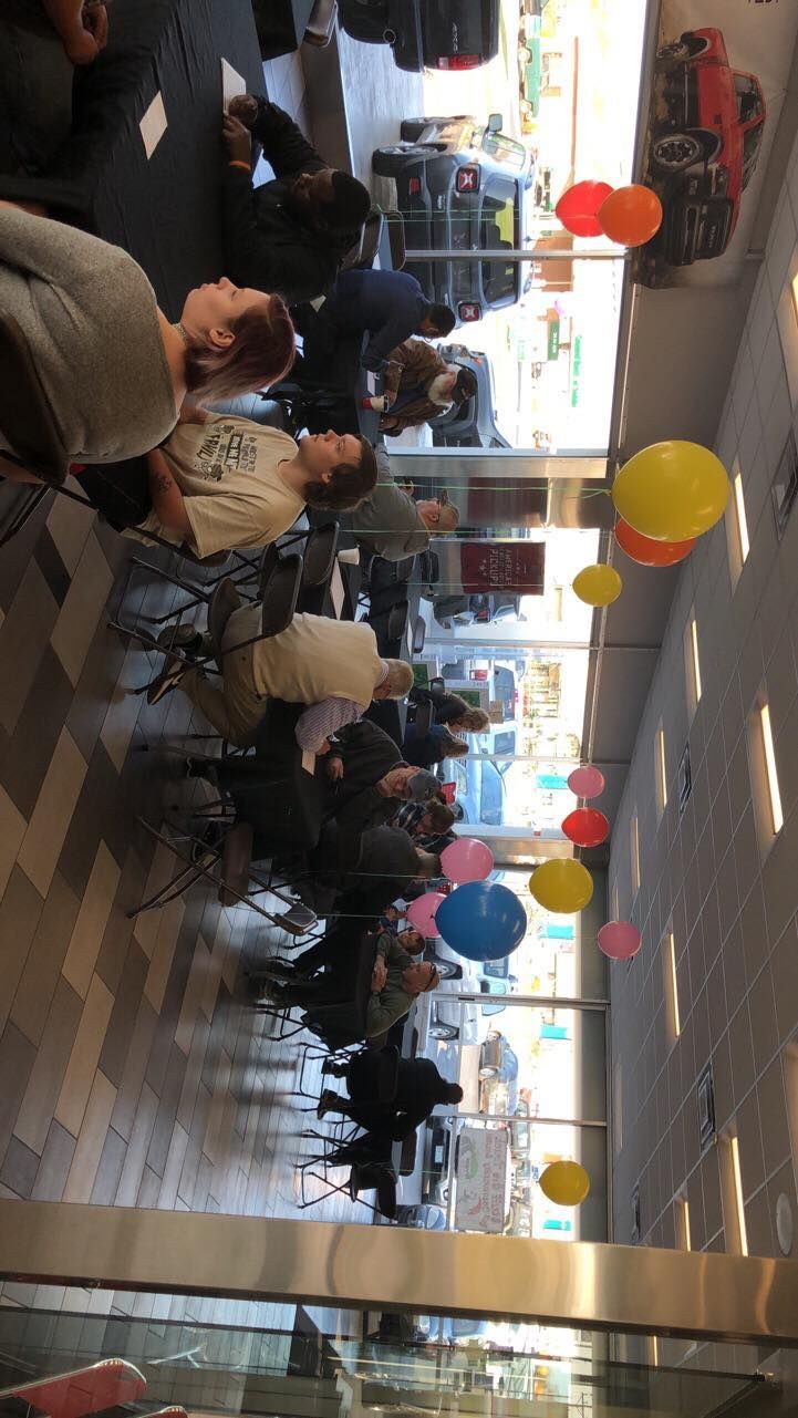 Crowded indoor event space with people seated at tables, colorful balloons, and large windows at the back