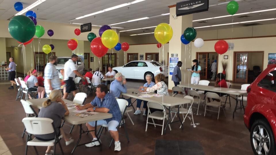 People seated at tables in a car dealership showroom decorated with colorful balloons; cars on display nearby.