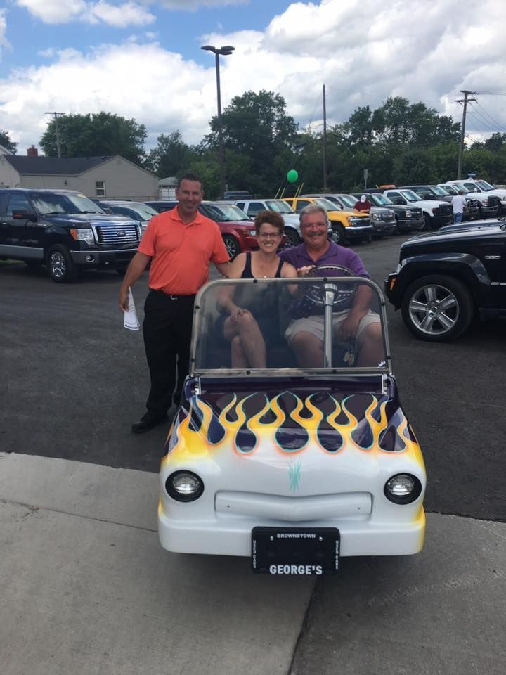Three people posing in a small white cart with flame decals in a parking lot