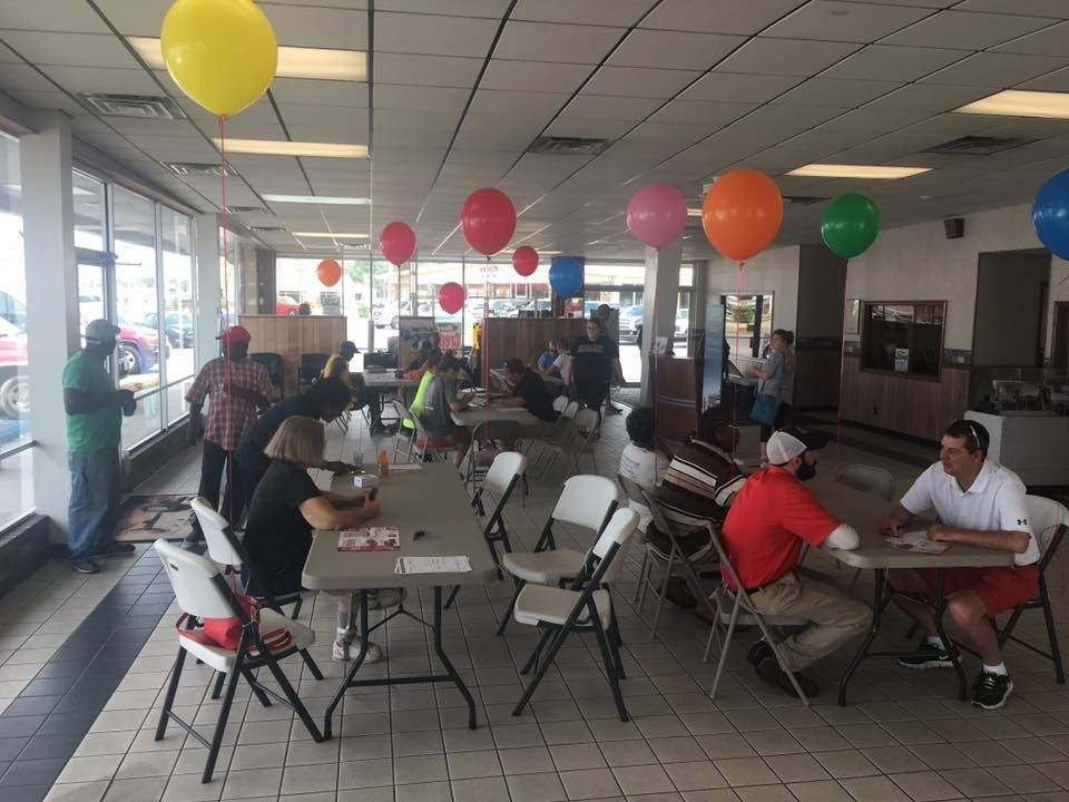 Community hall with people seated at tables, balloons overhead, and a casual indoor gathering