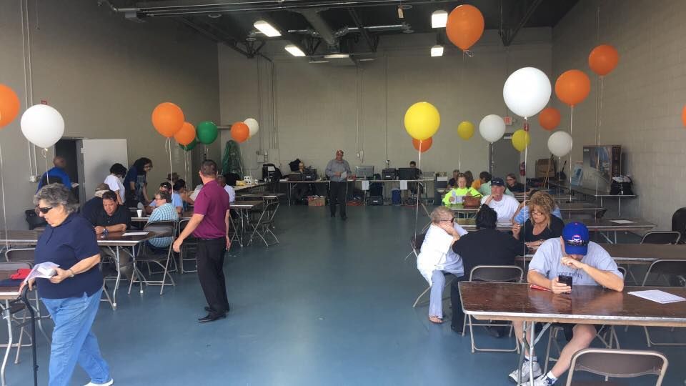 Indoor event with tables and balloons, people seated and mingling in a large warehouse-like room