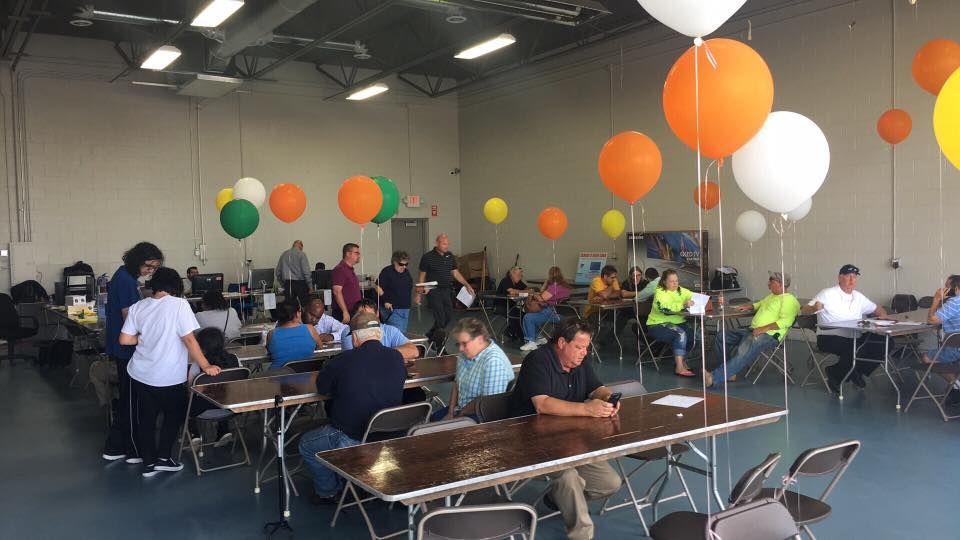 People seated at tables in a large indoor event space with orange, white, and green balloons overhead