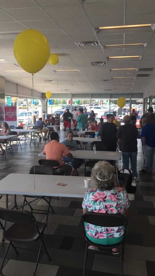 Crowded banquet hall with people seated at long tables under yellow balloons and fluorescent lights