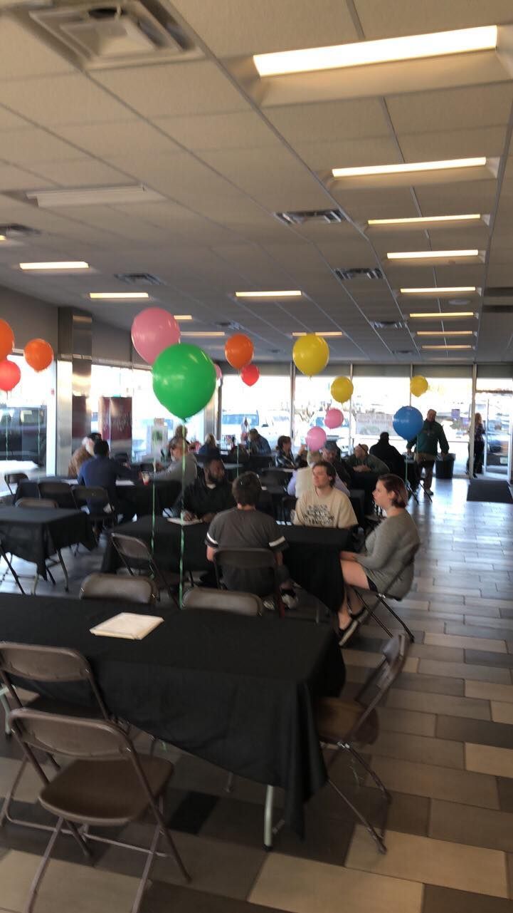 Indoor event hall with people seated at tables, colorful balloons, and bright overhead lighting