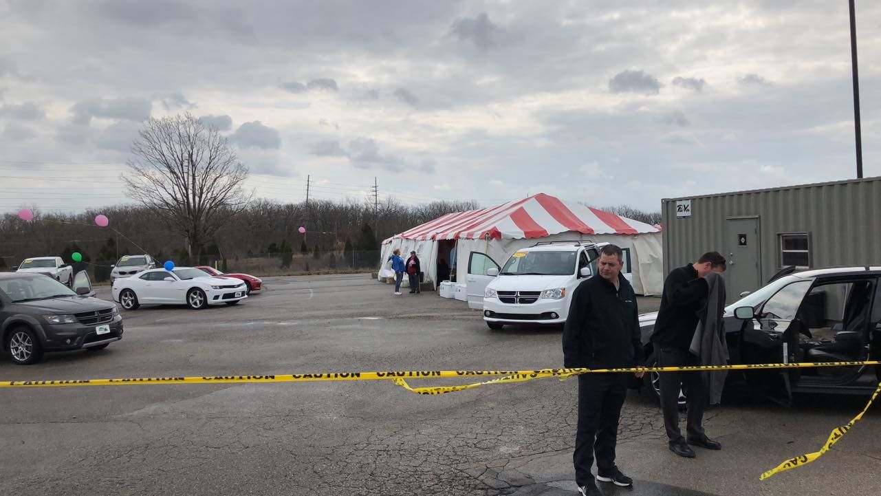 Police tape blocks a parking lot with cars, a white van, and a red-and-white tent under cloudy skies
