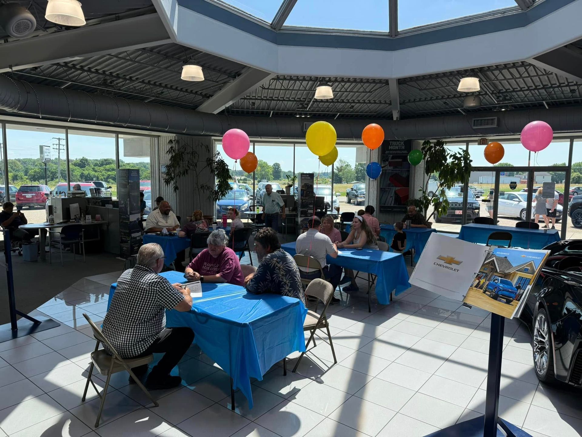 People seated at tables with blue tablecloths in a bright indoor restaurant, with colorful balloons overhead.