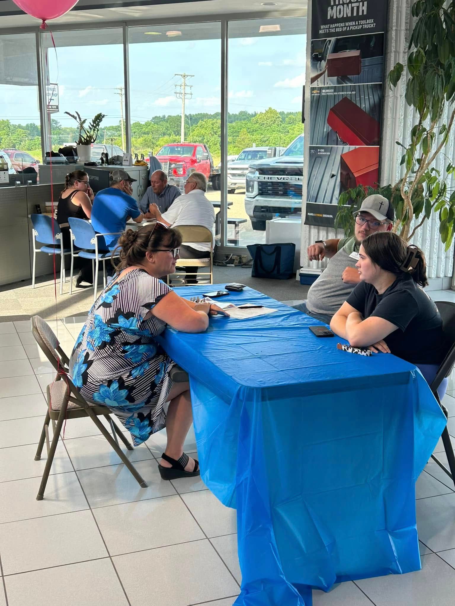 Two women sit at a blue tablecloth-covered table inside a storefront cafe, writing paperwork.