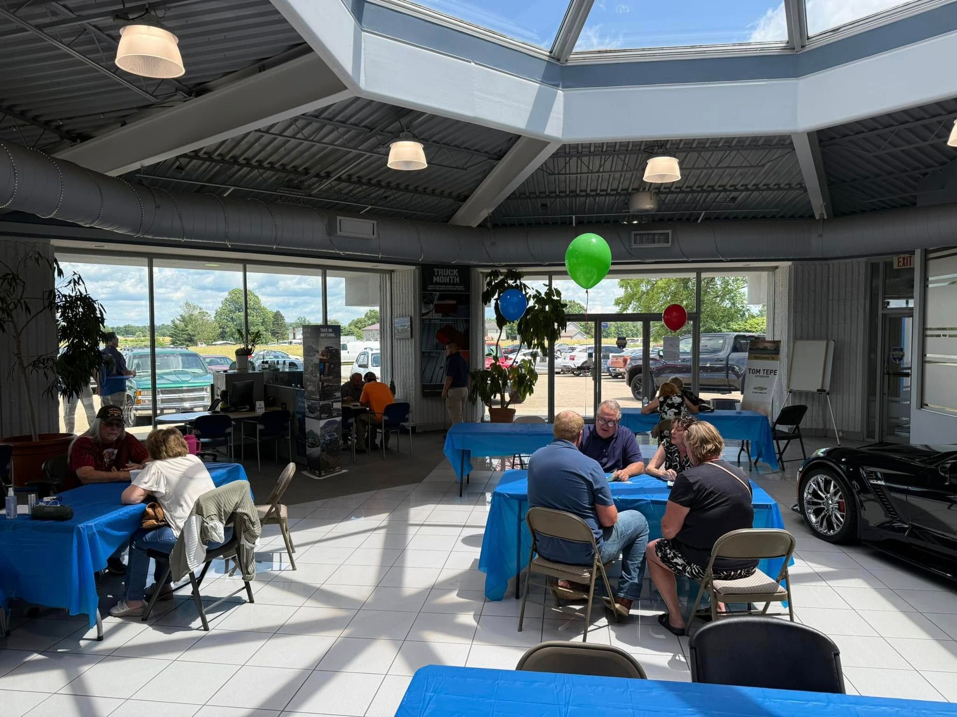 People seated at blue tables in a bright indoor atrium with skylight, balloons, and large windows.