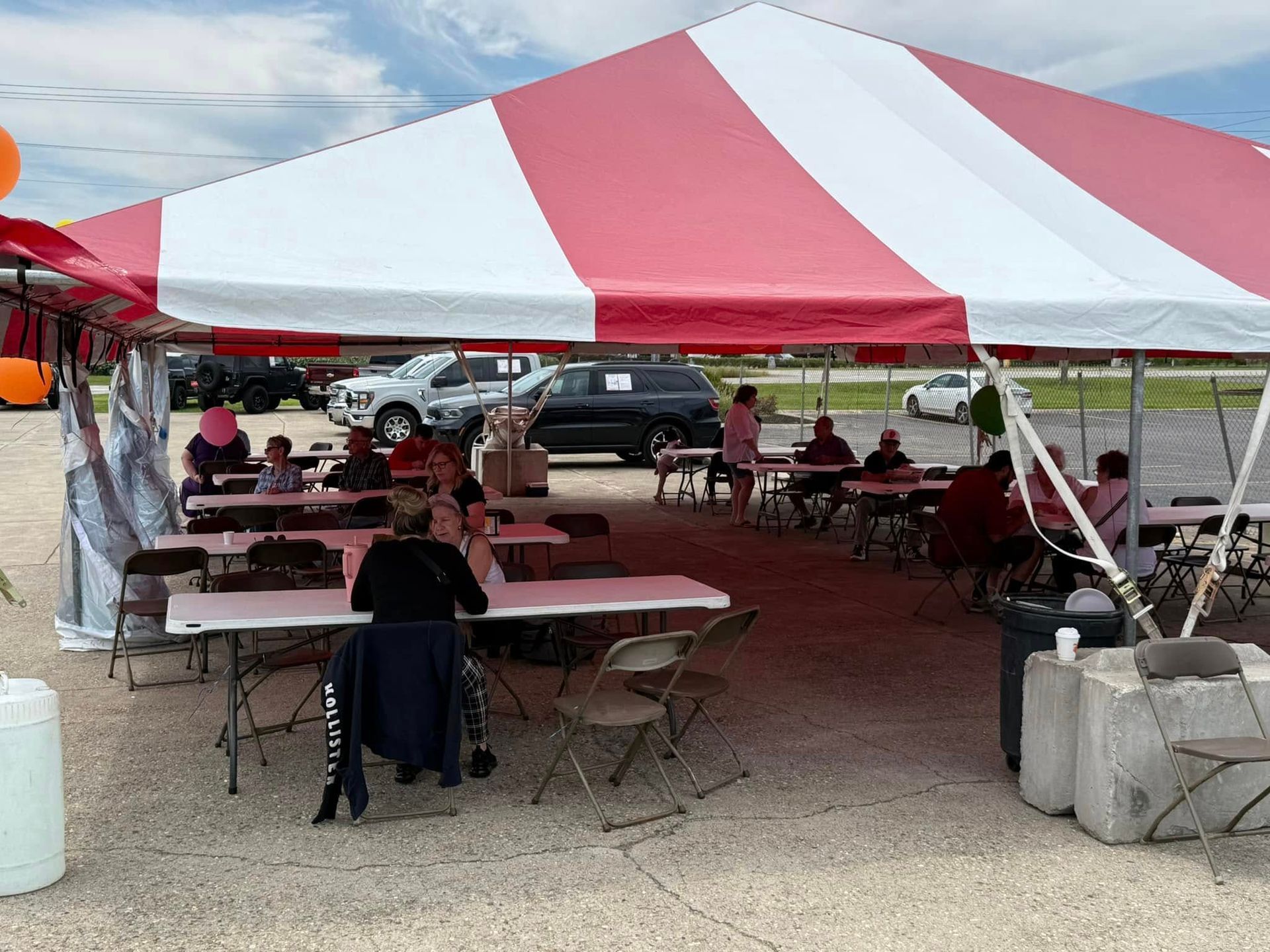 People seated under a red-and-white striped tent at an outdoor gathering with tables and chairs.