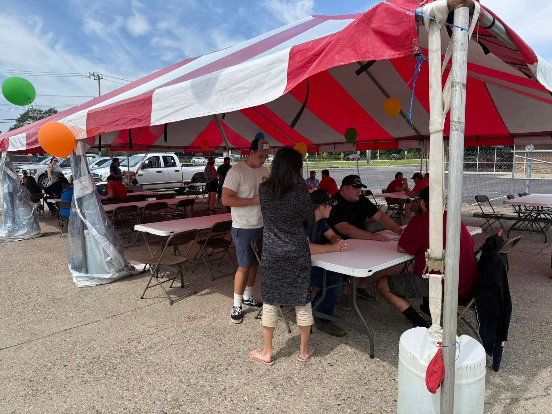 People seated under a red-and-white striped tent at an outdoor event, with tables and coolers on gravel.