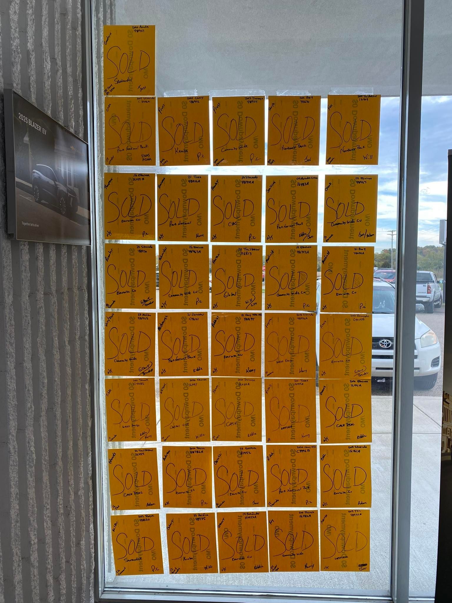 Orange-speckled paper grid taped to a window, with a dark screen on the left and parking lot outside.