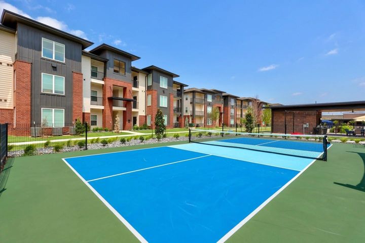 Outdoor pickleball court with blue and green surface in front of an apartment building on a sunny day.