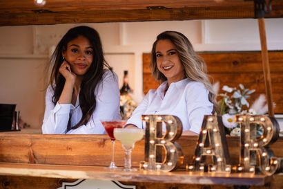 Two women behind a wooden bar with lit B-A-R letters and drinks on the counter