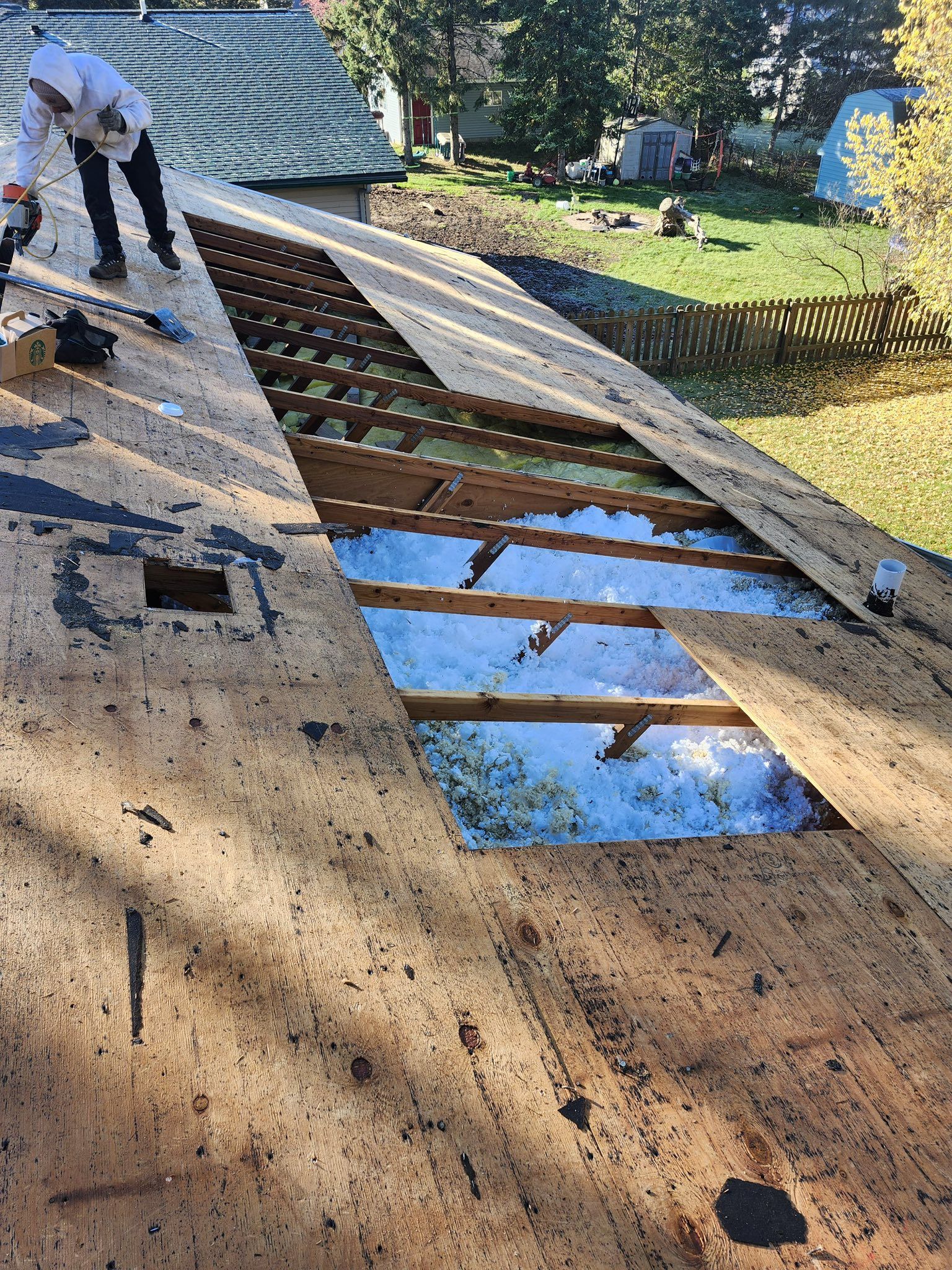 A person in white work clothes repairs a residential roof, with a large section of plywood missing to reveal the rafters.