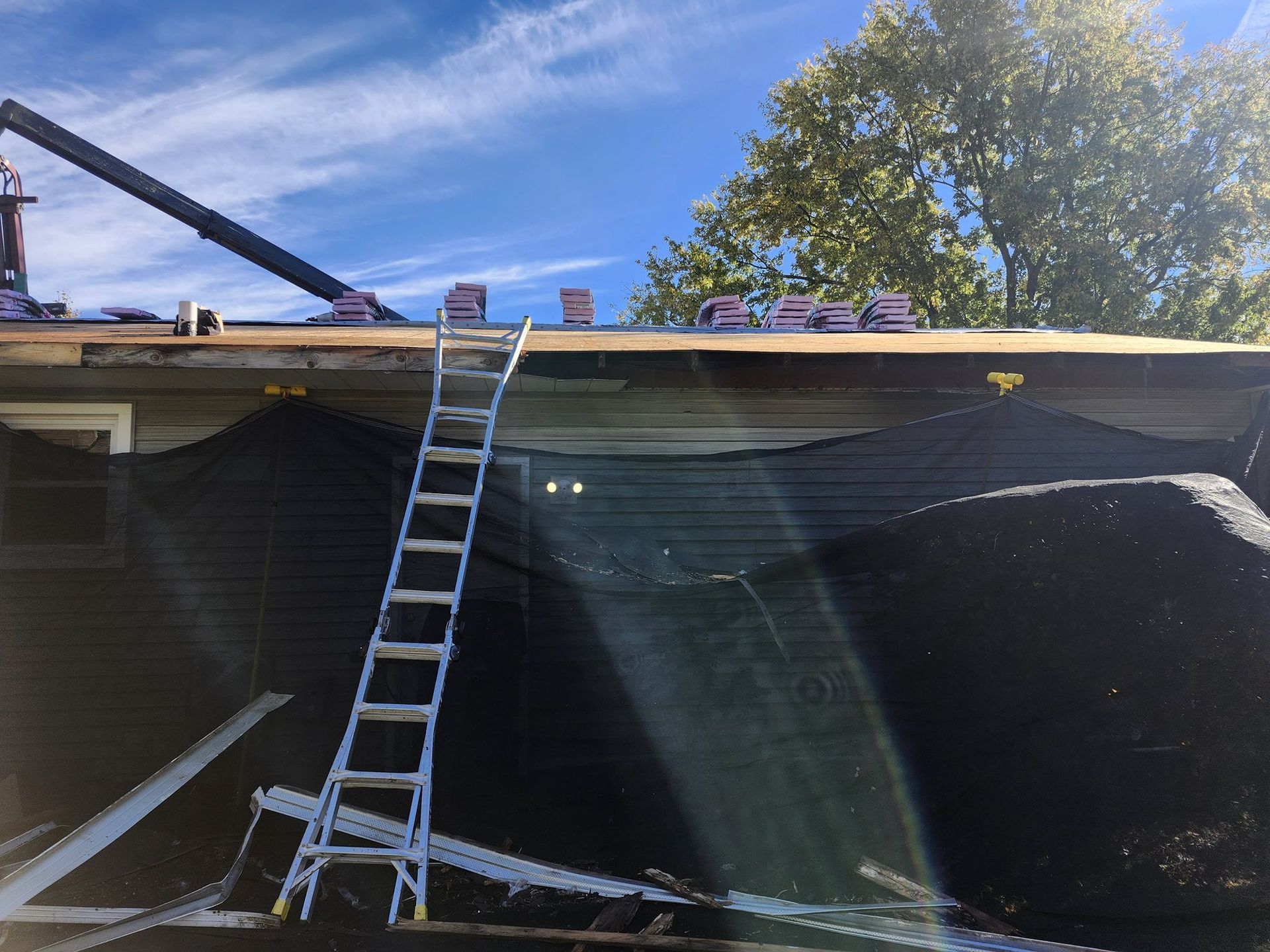 A metal extension ladder leans against a house during a roof renovation with construction debris netting below.