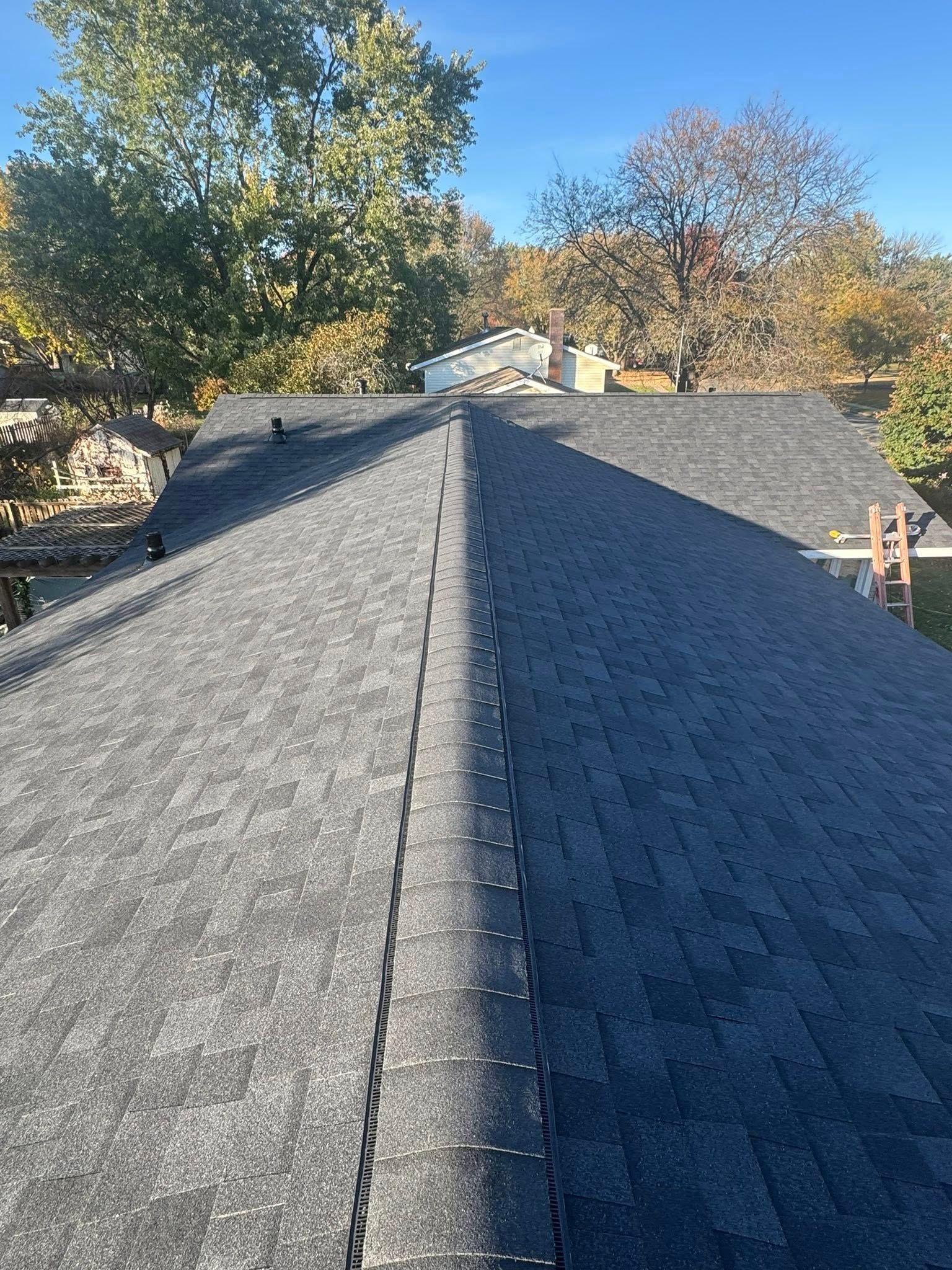 A high-angle view looking down the gray shingled ridge of a residential roof under a bright blue sky.