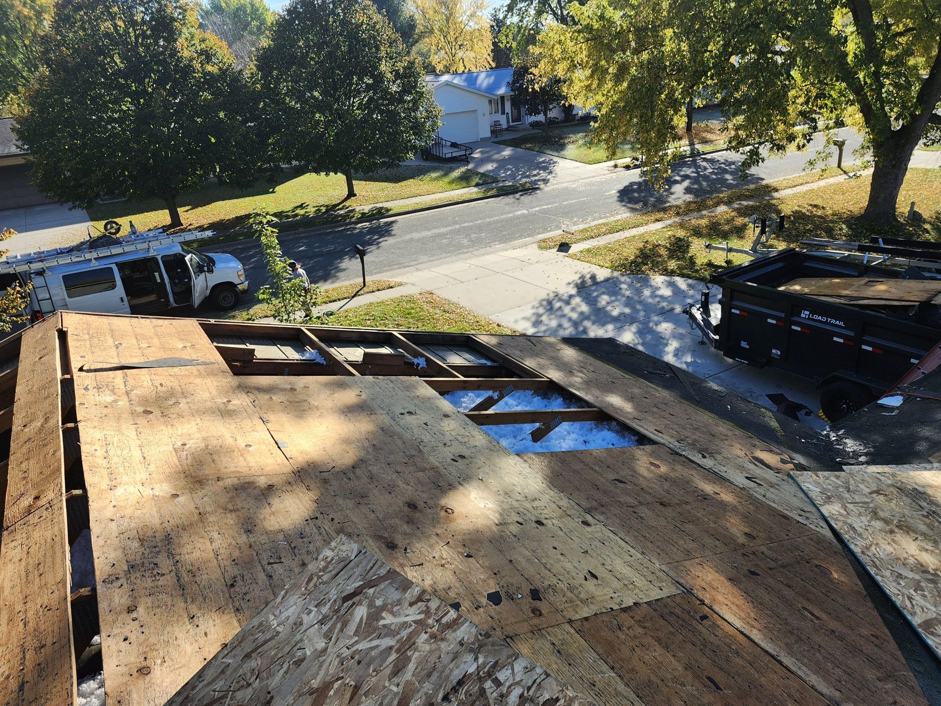 A partially stripped residential roof showing exposed wooden rafters and new plywood decking, with a dump trailer nearby.