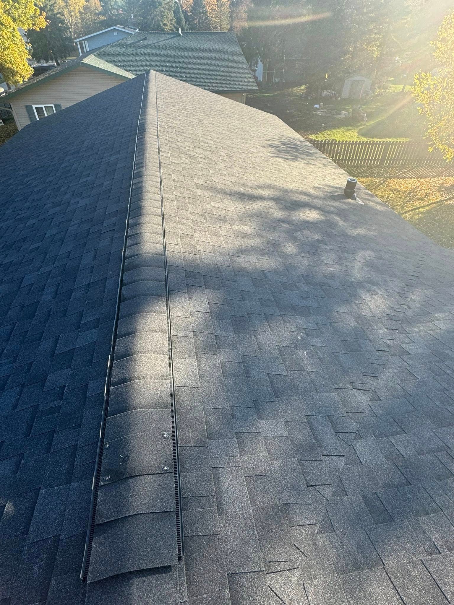 A view looking down the peak of a gray shingled roof under bright sunlight with trees in the background.