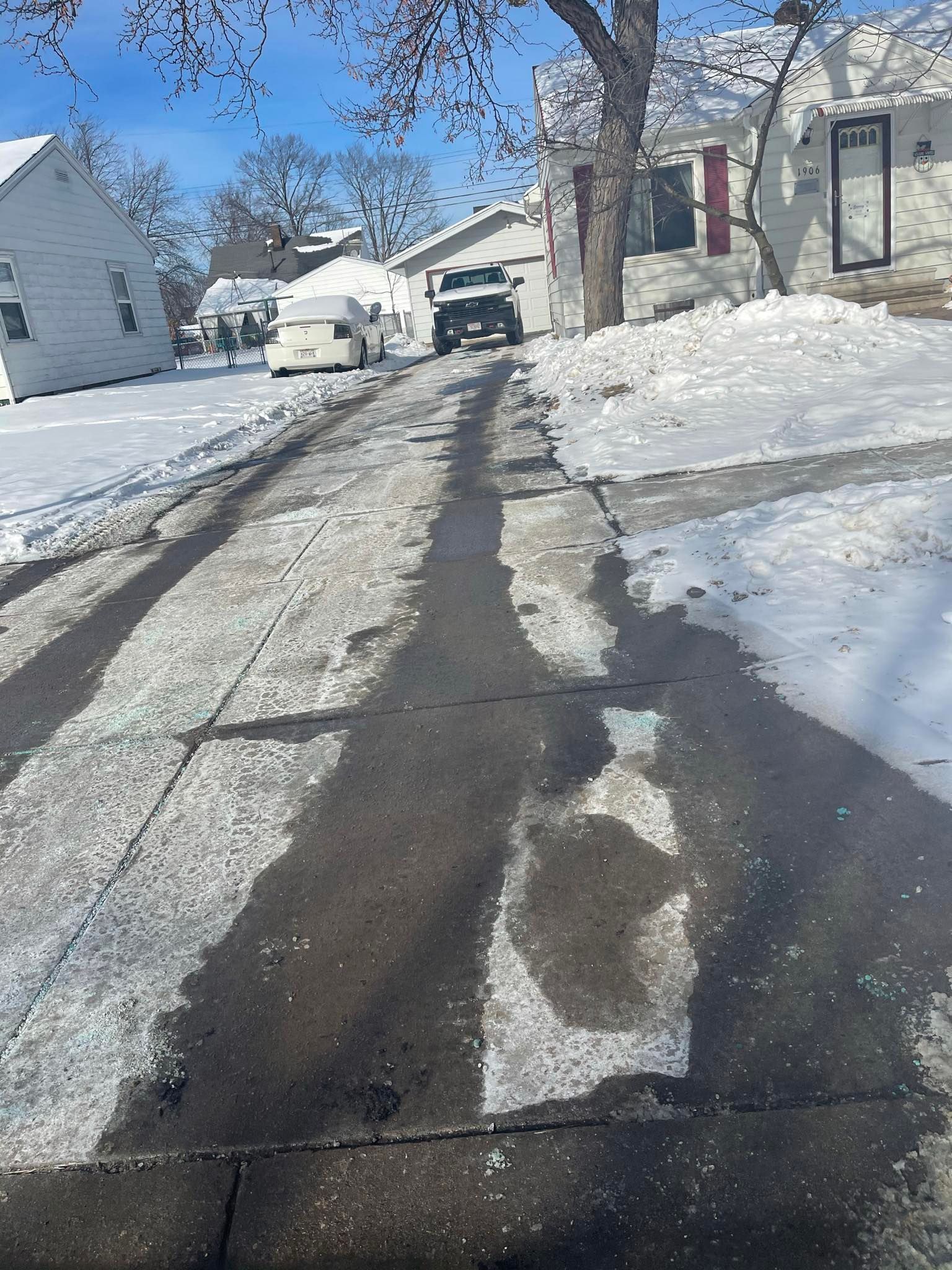A snow-covered residential driveway with cleared vehicle tracks leading to a house in a wintry neighborhood.
