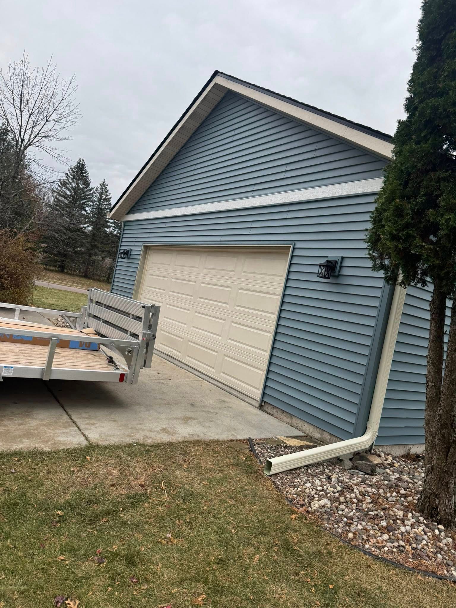 A detached, blue-sided garage with a cream-colored door sits on a concrete driveway next to a metal trailer.