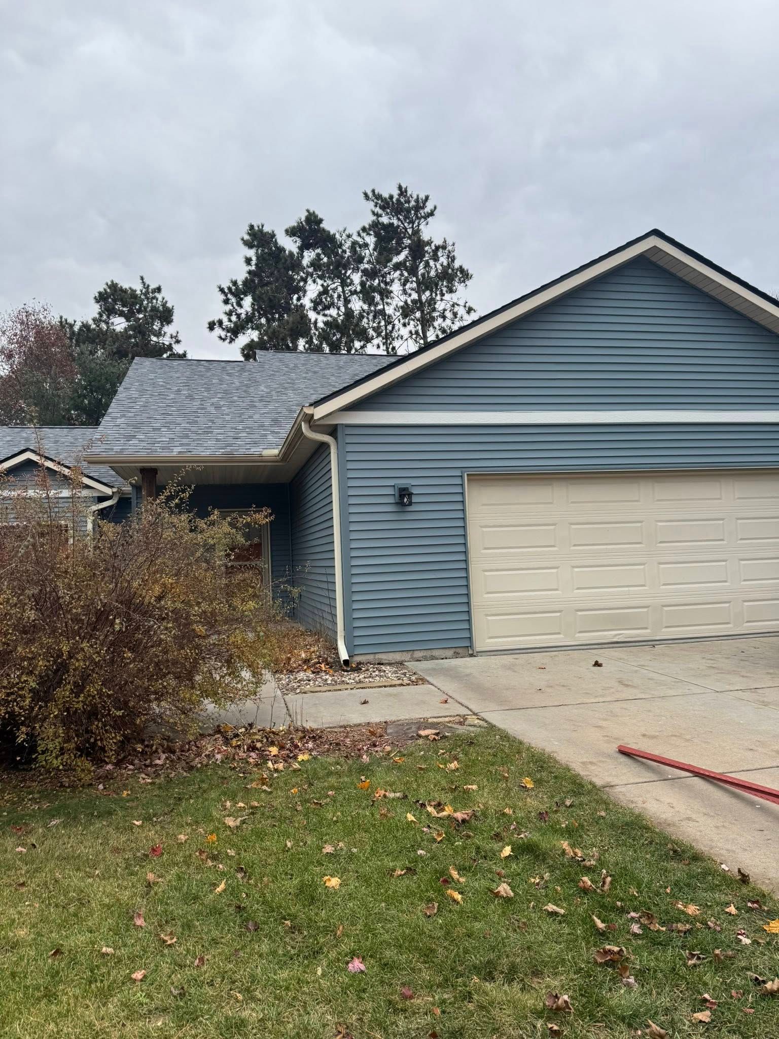 A single-story blue house with a gray shingled roof, two-car garage, and a large bush in the front yard on an overcast day.