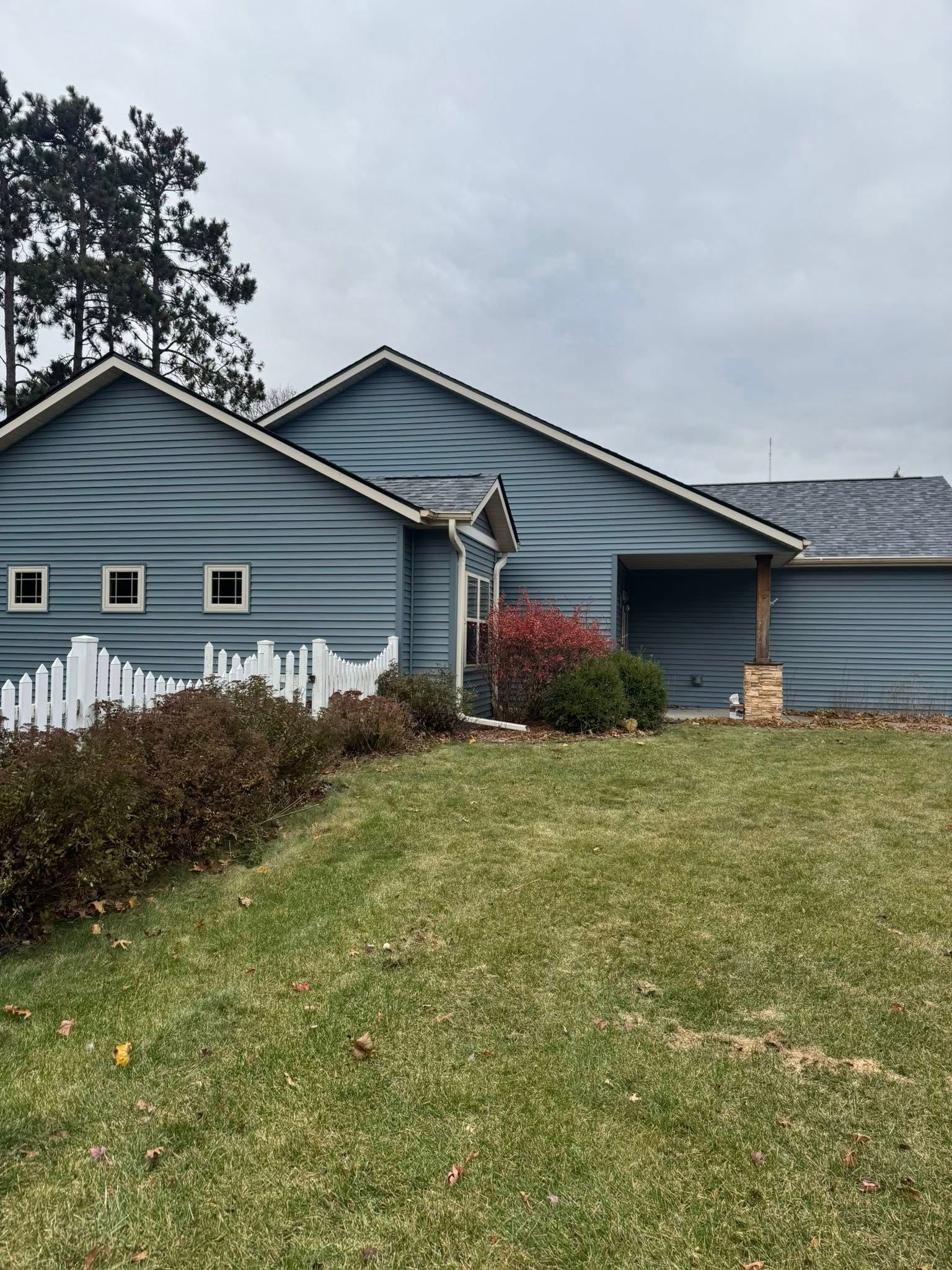 A blue log cabin-style house with a white fence, nestled in a yard with shrubs under a cloudy sky.