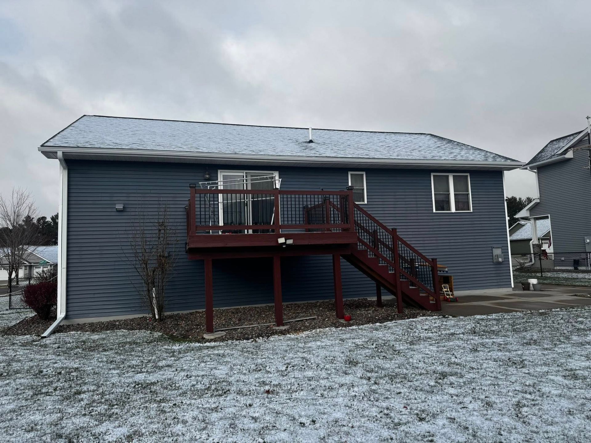 A gray siding house with a wooden deck and staircase, set against a snow-dusted lawn under a cloudy sky.
