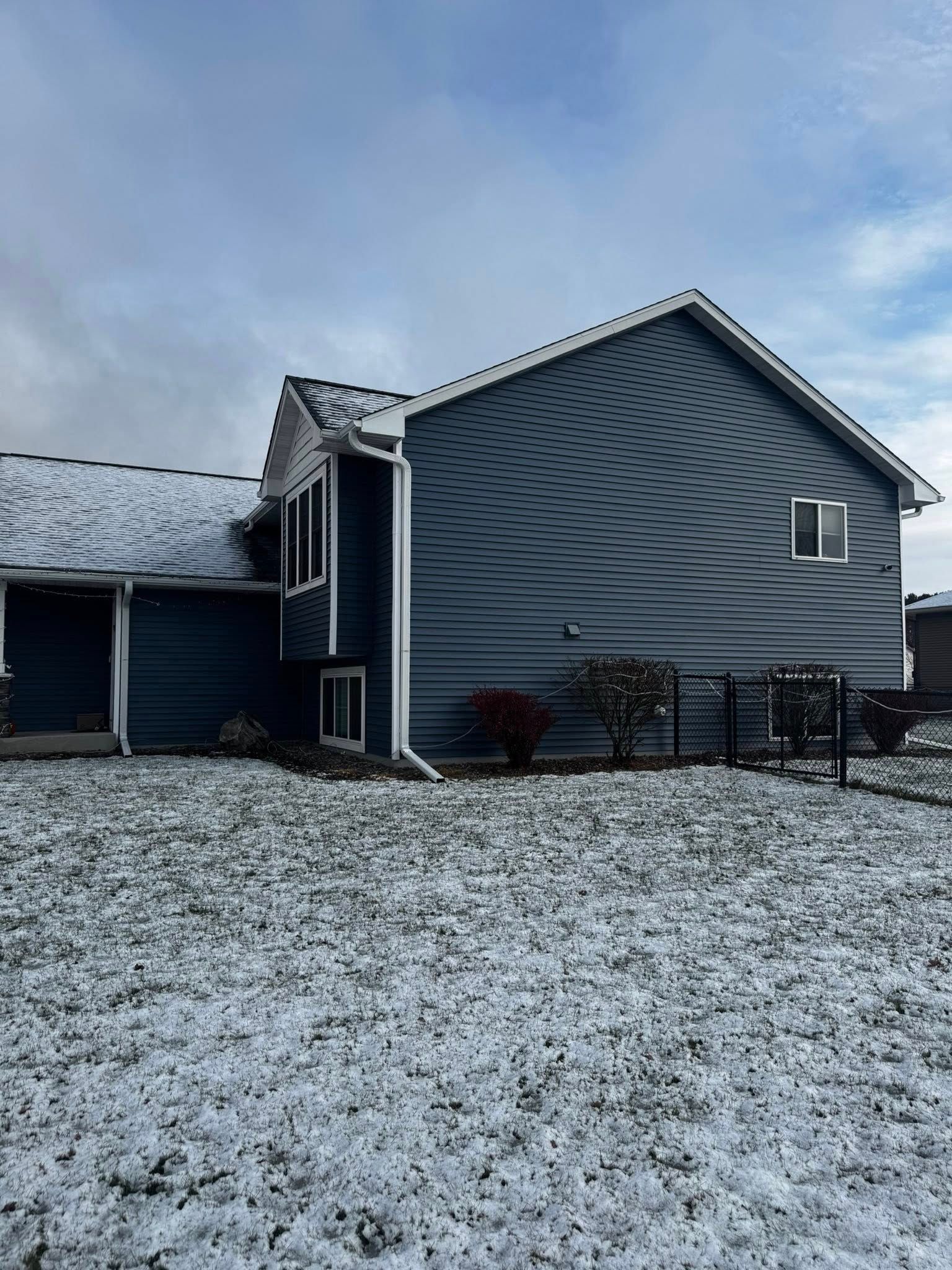 A two-story blue-sided house with white trim, partially covered in snow, stands on a snowy yard under a cloudy sky.