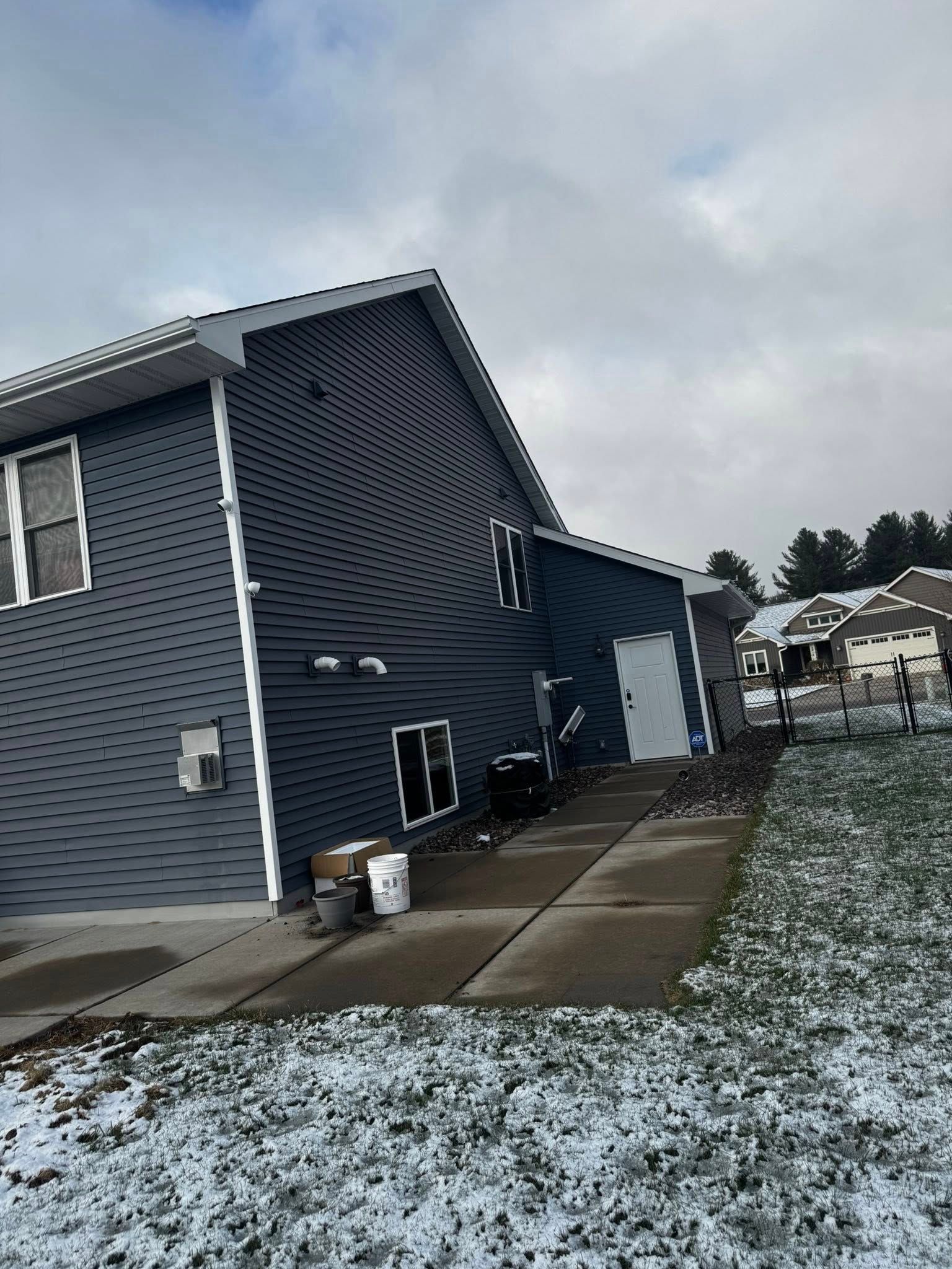 Side view of a dark blue, vinyl-sided house with a white door, concrete walkway, and snow on the ground.