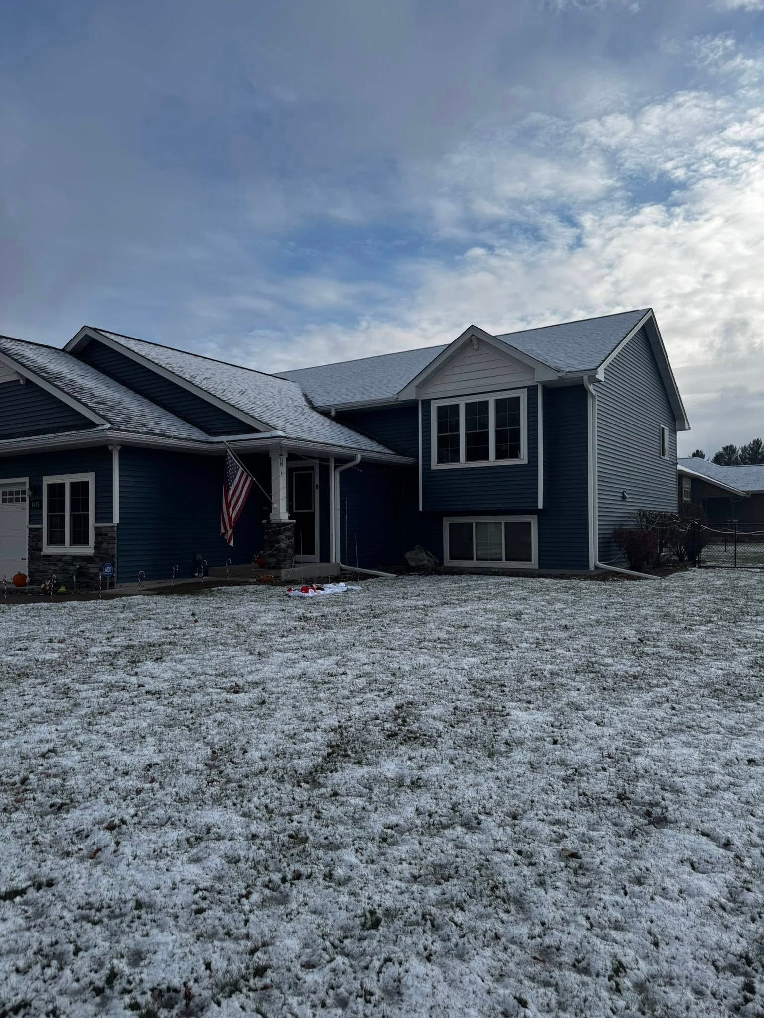 A dark blue, two-story house with white trim covered in light snow under a cloudy, bright sky.