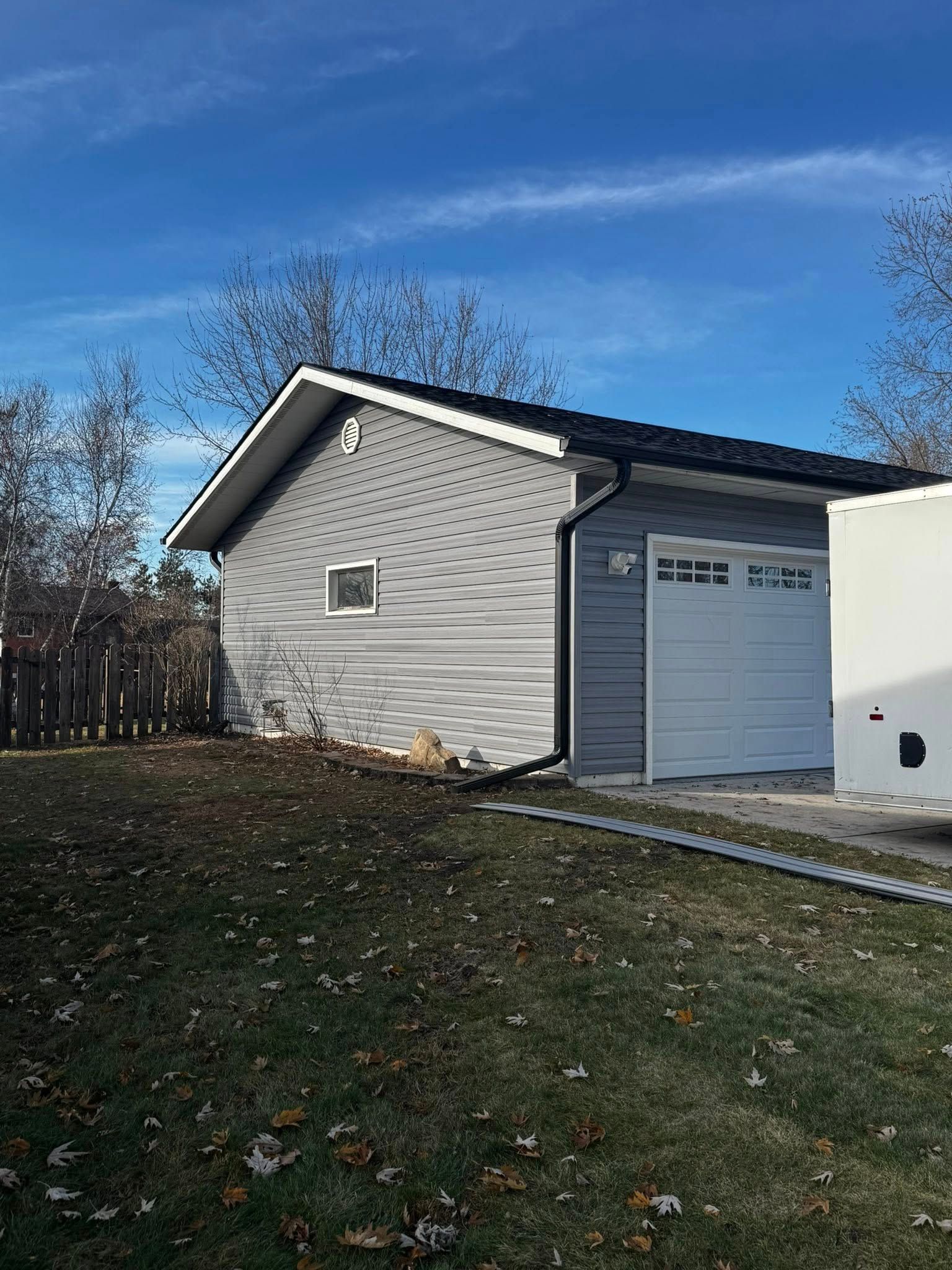 A gray detached garage with a white door and black trim stands in a yard under a bright blue sky.