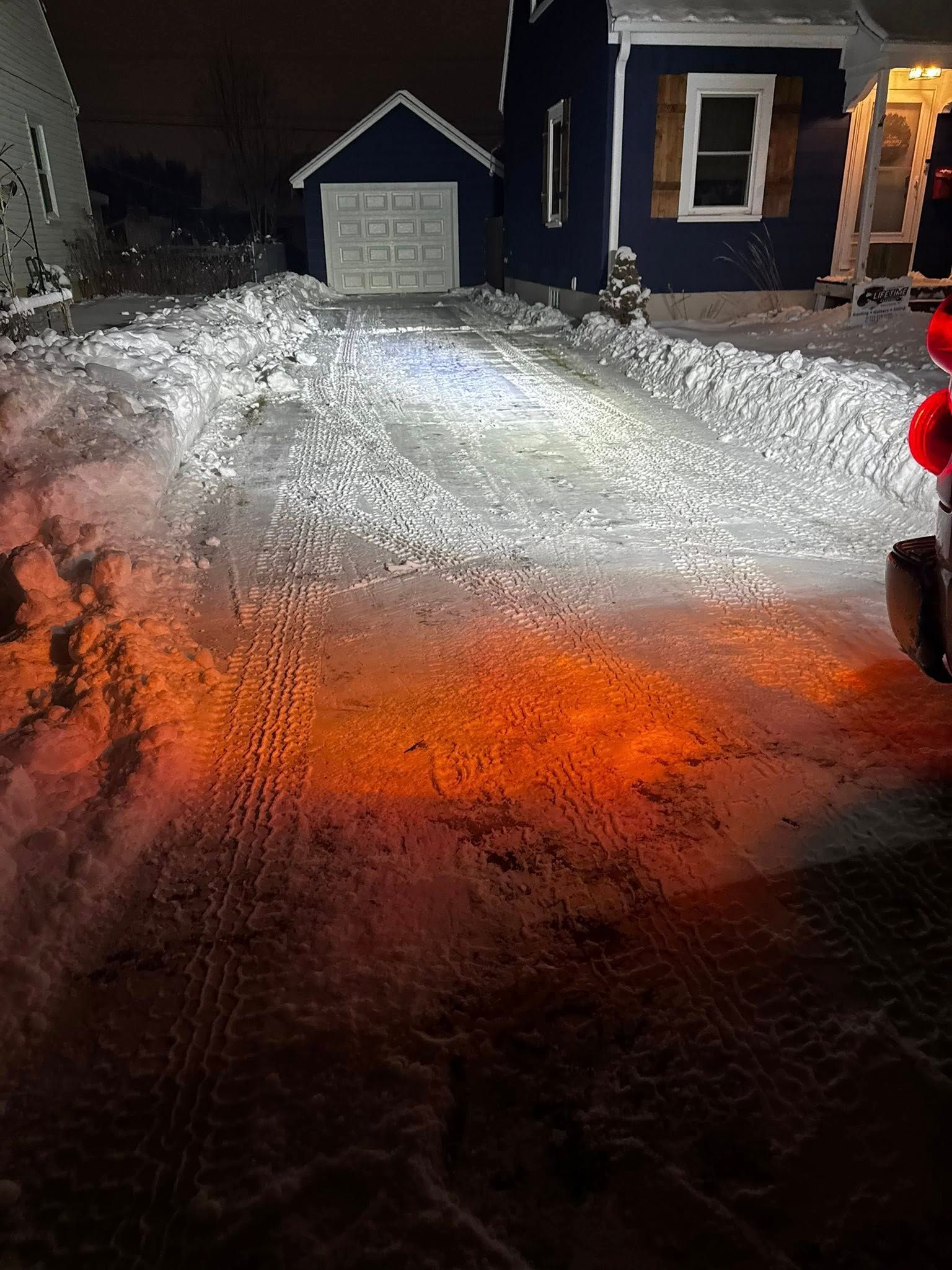 A dark snowy driveway at night, illuminated by bright white and orange vehicle lights, leading to a house and garage.