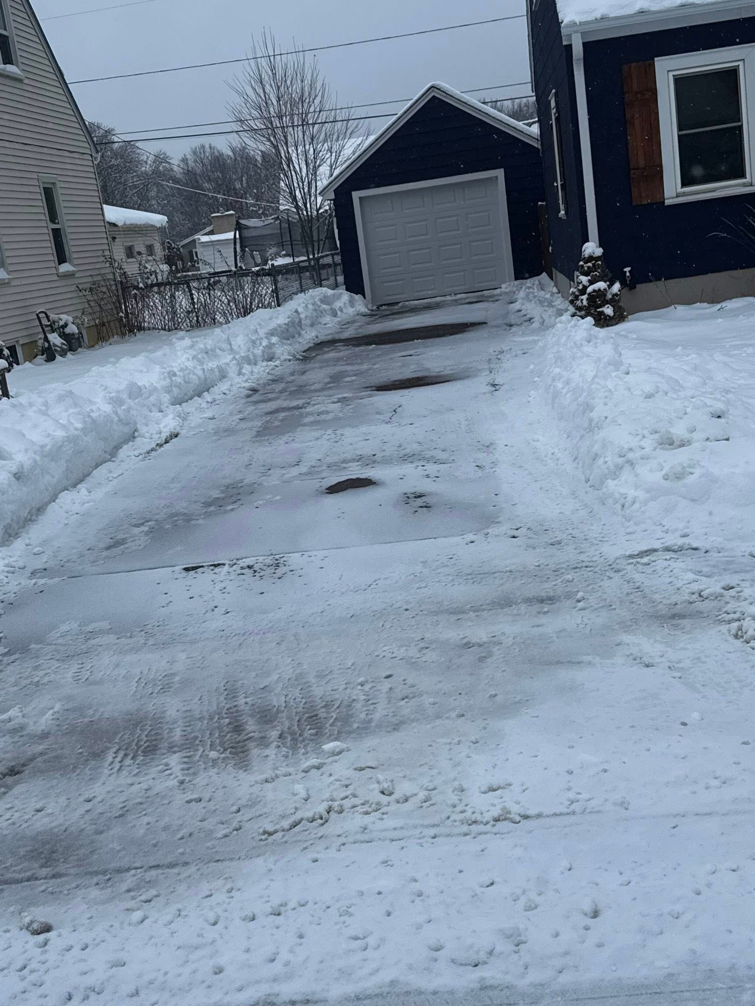 A snow-covered driveway leads to a dark-colored garage between two houses on a gray, overcast day.