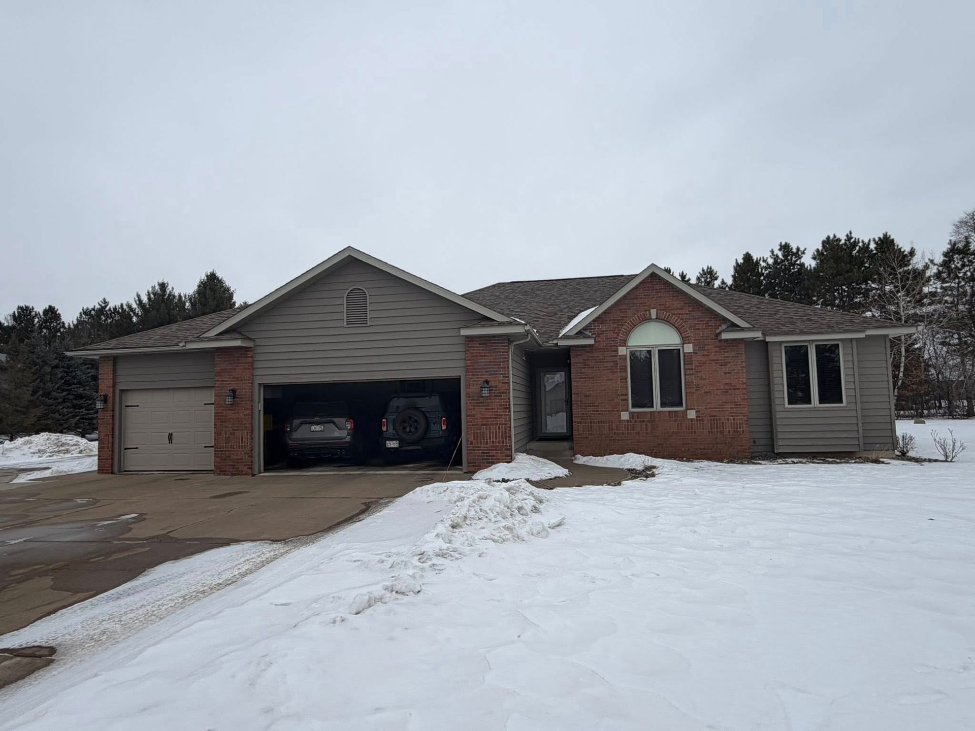 A single-story suburban house with a brick and siding exterior and a two-car garage sits on a snowy, overcast day.