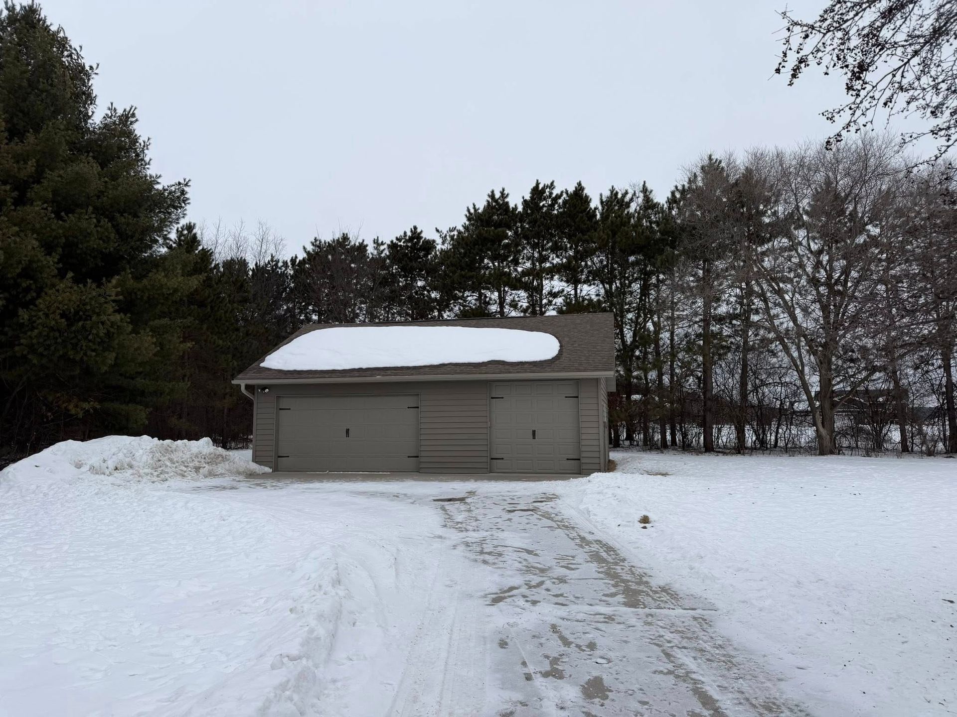 A gray two-car detached garage sits in a snow-covered yard surrounded by trees under an overcast sky.