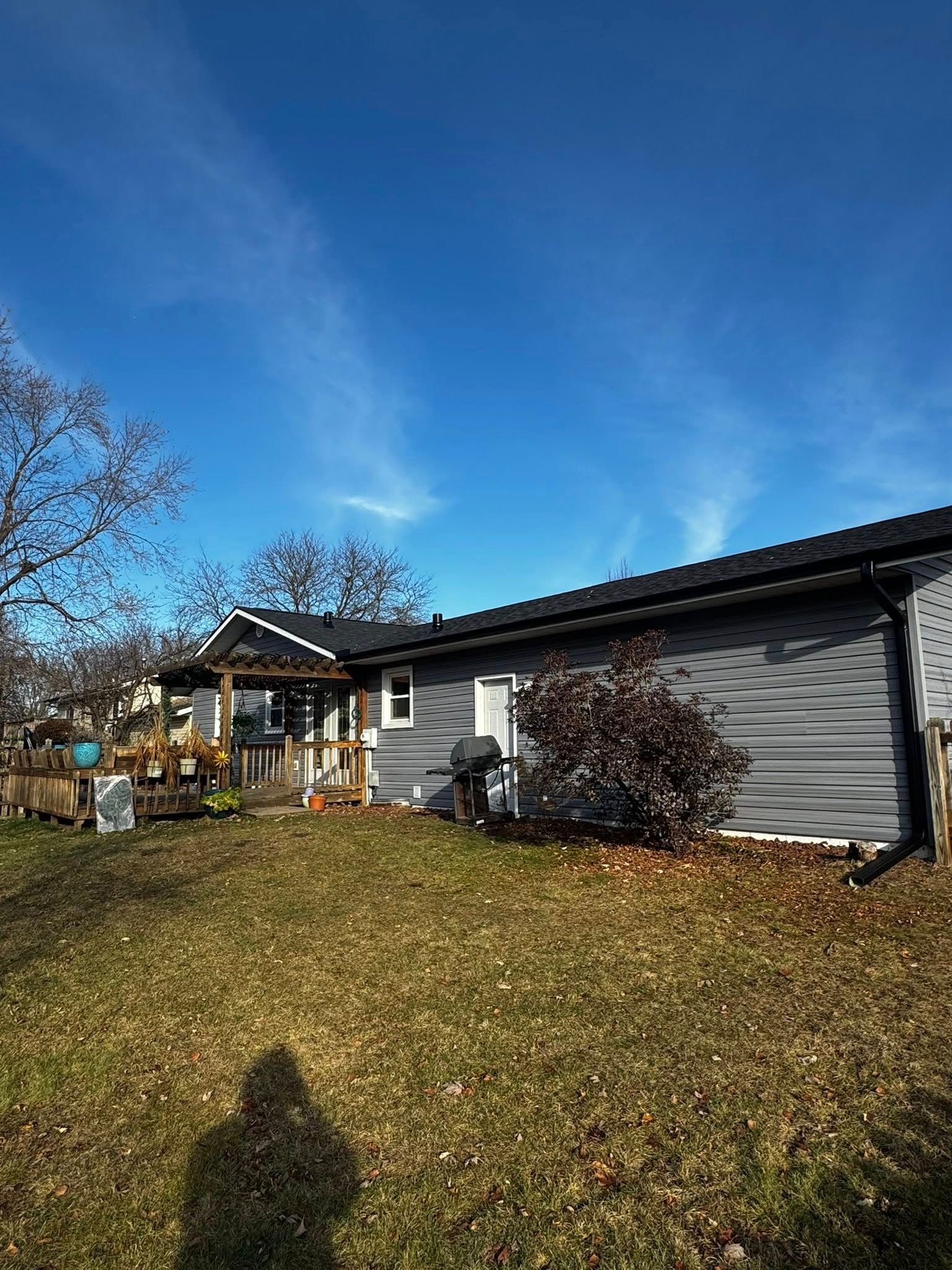 A gray house with a wooden deck and porch stands under a clear blue sky, viewed from a grassy backyard.
