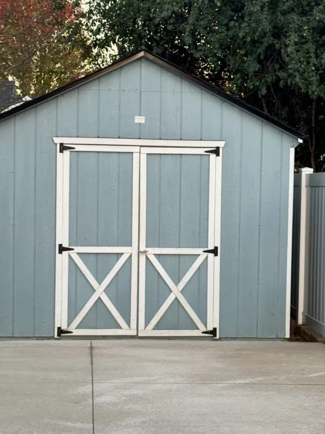 A light blue backyard storage shed with white double doors featuring X-shaped trim, standing on a concrete patio.