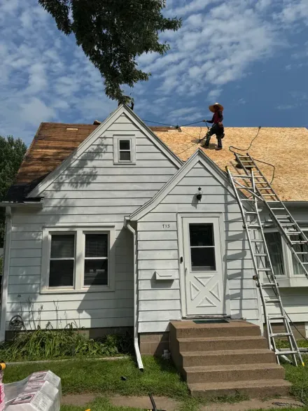 A person on a ladder and roof working on a home renovation, stripping old shingles to reveal the wood sheathing.