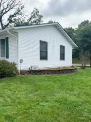 Side view of a white vinyl-sided house with a low, curved stone retaining wall in front of it on a grassy lawn.